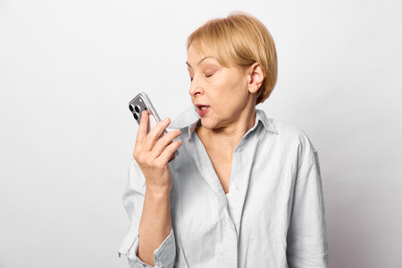 Middle aged woman in casual shirt using smartphone voice assistant, speaking while looking at...