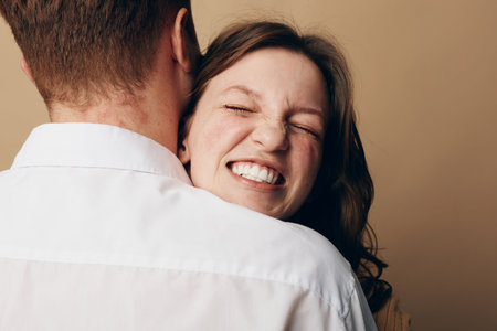 Happy woman with closed eyes smiles warmly while hugging man in white shirt. Intimate moment of...