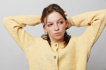 Young woman in yellow knitted sweater stretches with hands behind head, looking sideways. Casual...