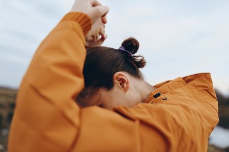 Woman in orange jacket ties her hair outdoors, preparing for activity. The cloudy sky and blurred...