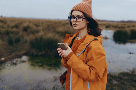Young woman in orange jacket and knitted hat stands outdoors near water and grasslands holding...