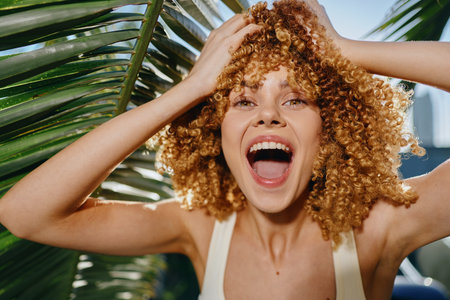smiling woman with curly hair enjoying sunny day outdoors surrounded by green palm leaves, natural...
