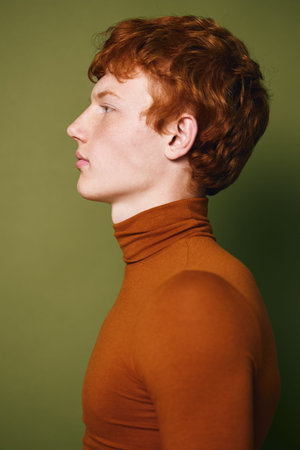 Profile of young man with red hair wearing brown turtleneck sweater against green background with...