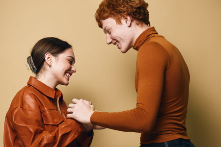 Young couple holding hands and smiling, facing each other with affection against beige background....