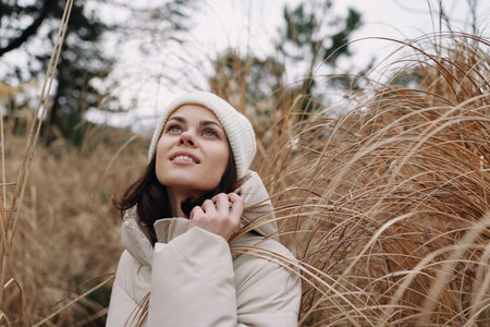 Woman in a light coat and knit beanie stands among tall autumn grass, looking up with a gentle...