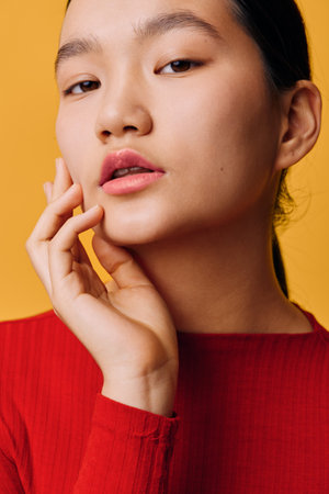 Young woman with natural makeup posing with fingers near face looking at camera on yellow background. Female model wearing red top with smooth skin and parted lips in studio lighting.の写真素材