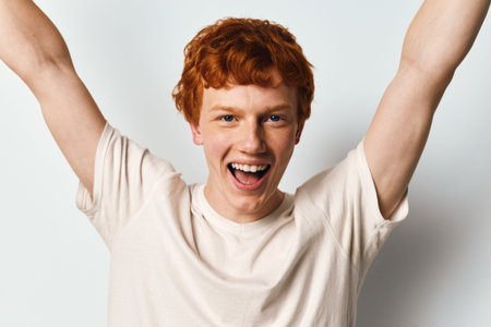 Young man with red hair wearing white t shirt raising arms and smiling broadly against white background for celebration and happiness concept.の写真素材