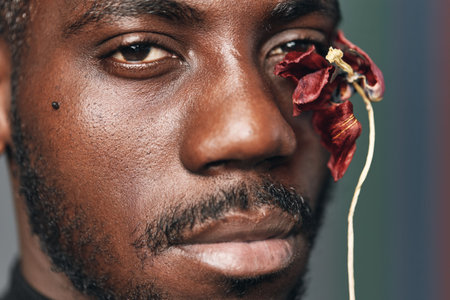 African man portrait with dark skin and facial hair holding dried red flower near eye in creative...