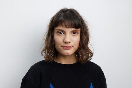 Portrait of a young woman with dark hair and a calm expression, wearing a black sweater against a light, neutral backdrop. Soft lighting highlights natural features and a relaxed, approachable mood.の写真素材