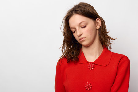 Sad young woman in red sweater looks down with closed eyes on white background. Concept of...