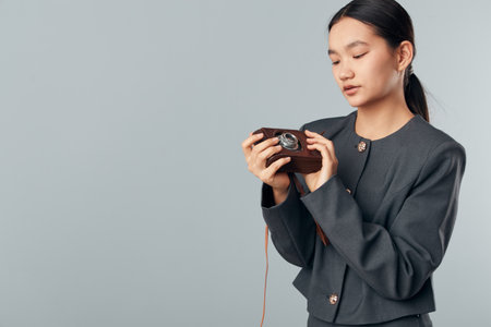 Young woman wearing gray jacket holding vintage camera with focus at light gray background. Female photographer inspecting old camera for hobby or photo shoot with calm expression indoors.の写真素材