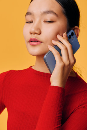 Woman in red sweater talking on smartphone with eyes closed against yellow background. Portrait of young female holding mobile phone for communication and connection.の写真素材