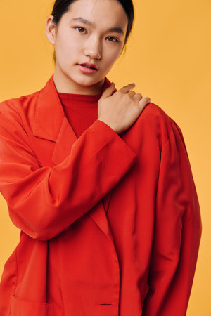 Portrait of young woman in red coat posing on yellow background with hand on shoulder, looking at...