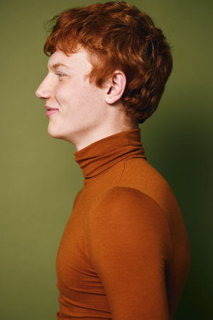 Portrait of smiling young man with red hair wearing orange turtleneck sweater against green background in studio. Profile view of happy person with fair skin and curly hair.の写真素材