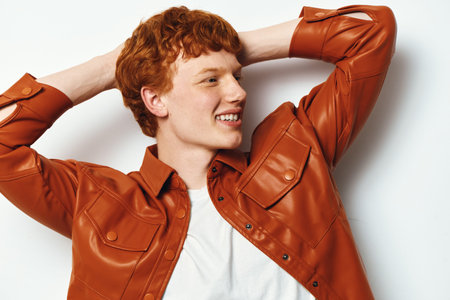 Young redhead man in orange leather jacket smiling and posing with hands behind head on white background. Casual fashion portrait, happy expression and natural look.の写真素材
