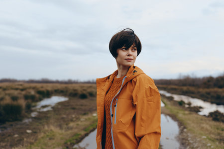 Young woman lifestyle standing outdoors in orange jacket on cloudy day. Natural environment with wet...
