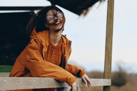 Happy woman lifestyle outdoors wearing glasses and orange jacket leans on wooden fence smiling with...