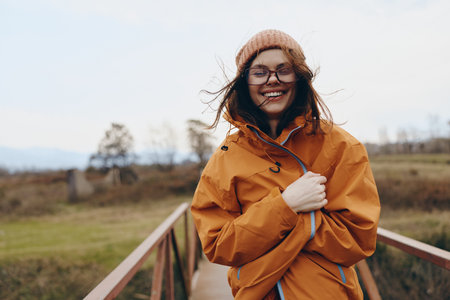 Smiling woman lifestyle outdoors wearing orange jacket and glasses stands on wooden bridge in autumn...