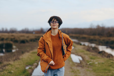 Young woman lifestyle walking outdoors in autumn wearing orange jacket and sweater, glasses, casual outfit. Concept of freedom, nature, and casual fashion in cool weather.の写真素材