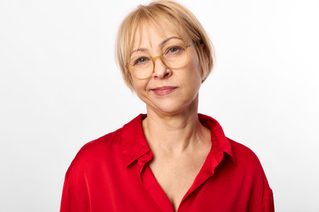 Mature woman with short blonde hair and glasses smiles gently wearing red blouse on white...