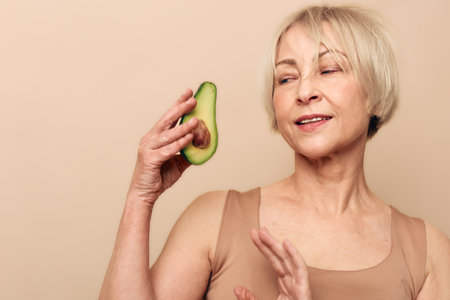 Mature woman with short blonde hair holds half avocado near face, smiling gently with closed eyes....
