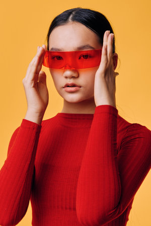 Young woman in red sweater wearing futuristic red sunglasses with hands adjusting eyewear on orange...
