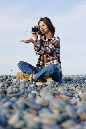 Woman photographer on a rocky beach, capturing moments with a camera as sand and pebbles fly, casual...