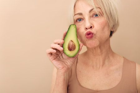 Mature woman with short blonde hair holds fresh avocado half near face, making playful kiss gesture....