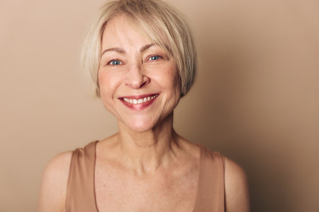 Smiling mature woman with short blonde hair looks directly at camera, natural makeup and warm beige...