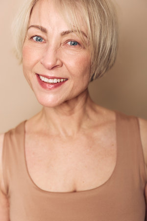 Smiling mature woman with short blonde hair and blue eyes wears beige top, showing natural skin and...