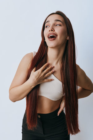 Young woman with long straight hair wearing white crop top and black pants posing with hand on chest and looking up with joyful expression on plain background.の写真素材