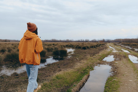 Woman lifestyle concept with a person wearing an orange jacket and beanie walking on a muddy rural path surrounded by fields under a cloudy sky. Outdoor autumn activity and nature.の写真素材