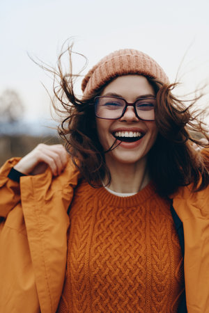 Smiling woman lifestyle wearing orange sweater and jacket with glasses and hat outdoors. Autumn mood...