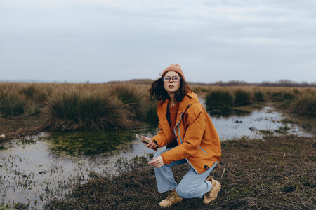 Young woman in orange jacket and hat crouches near water in natural landscape with grass and cloudy...