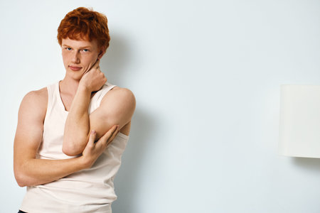Young man with red hair in white tank top standing near white wall looking at camera with thoughtful...
