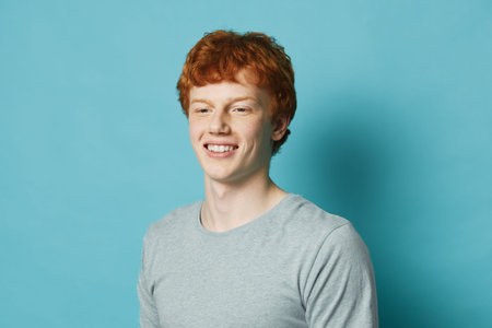 Young man with red hair smiling and wearing gray shirt standing against blue background. Portrait of happy male with casual look and cheerful expression in studio.の写真素材