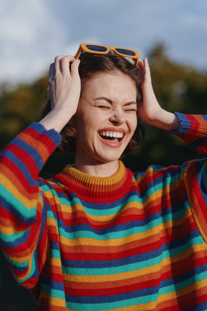 Smiling woman in rainbow sweater laughing outdoors, holding sunglasses on her head. Young female...