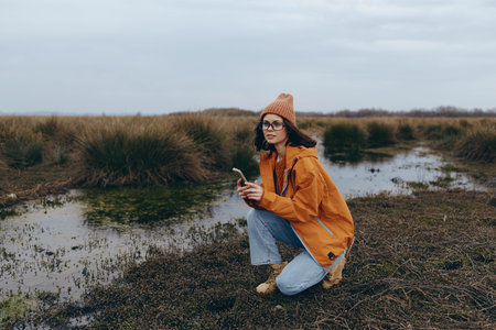 Young woman in orange jacket and knitted hat crouches on wetland grass holding smartphone. Outdoor...