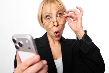 Surprised middle aged woman in glasses holds smartphone looking shocked with wide eyes and open...