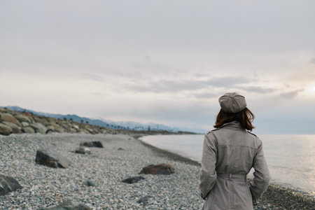 Woman in patterned coat and hat stands on rocky beach looking at calm sea under cloudy sky. Concept...