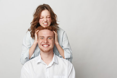 Playful young woman with curly hair squeezes head of smiling redhead man in white shirt. Studio...