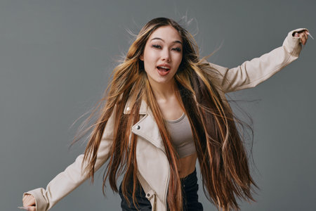 Young woman with long hair wearing jacket and tank top dancing in studio with grey background for...