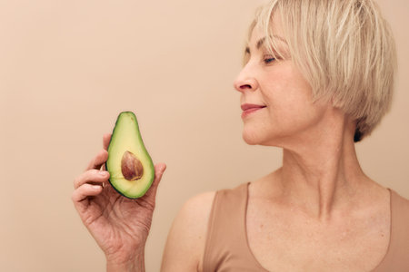 Mature woman with short blonde hair holds half an avocado close to her face on beige background....