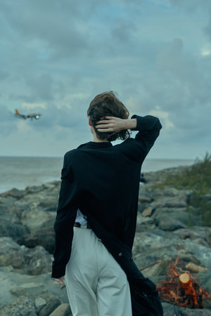 Coastal scene with an individual facing the sea on a rocky shore, wind in the backdrop, hand at the...