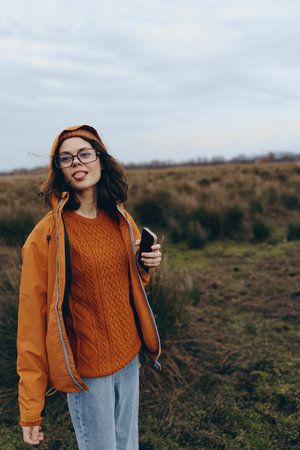Young woman lifestyle in orange jacket and sweater holds phone outdoors in autumn field. Casual...