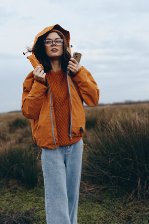 Young woman lifestyle wearing orange jacket and glasses stands outdoors in nature holding a phone....