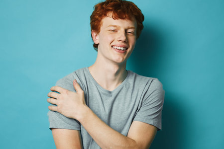 Young man with red hair and casual gray t shirt smiling with eyes closed against blue background....