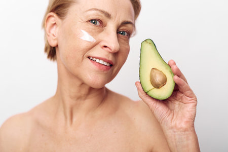 Mature woman with short hair holds half an avocado near her face with a smile. She has a small...