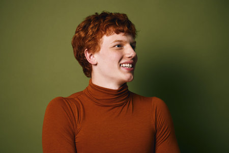Young redhead man smiling and looking sideways wearing brown turtleneck sweater on green background...