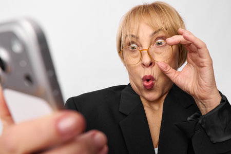 Surprised middle aged woman with glasses takes selfie with smartphone on white background. Concept...
